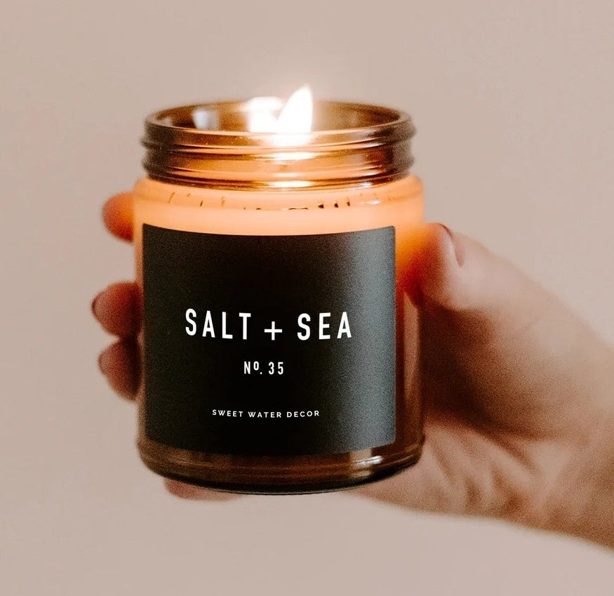 Hand holding a candle that says &quot;Salt + Sea&quot; on a black label