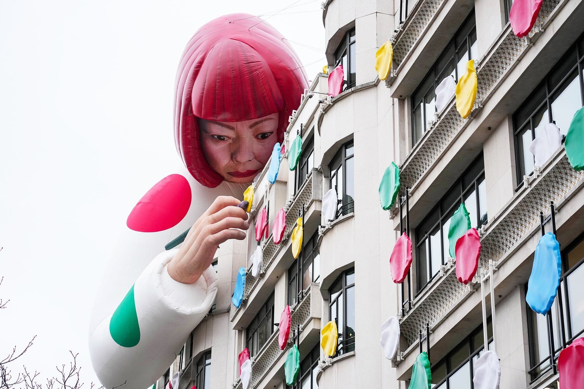 A gigantic statue of a woman in a pink bob peering into a building