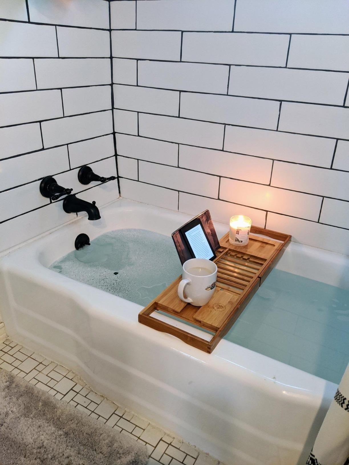 reviewer image of the caddy on their bathtub with a mug, a candle, and a kindle
