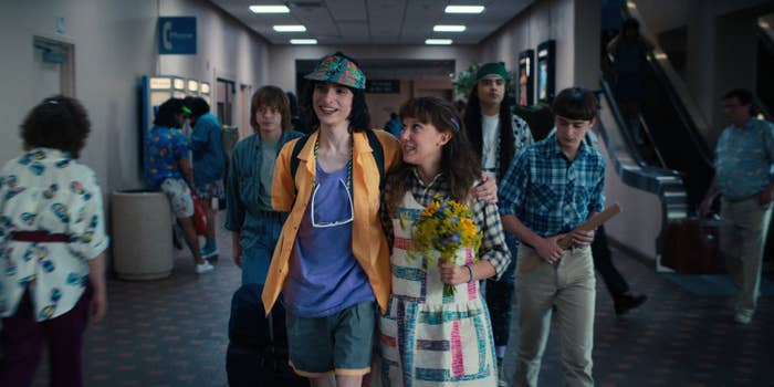 Finn, with his arm around Millie, as they walk through a hallway in a scene from Stranger Things