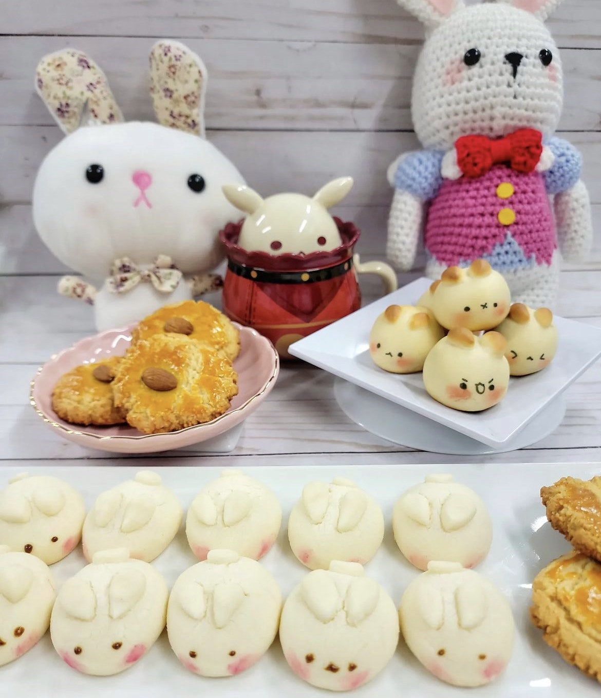 Bunny shaped cookies, breads, and eclairs on wooden white table next to plush bunnies.