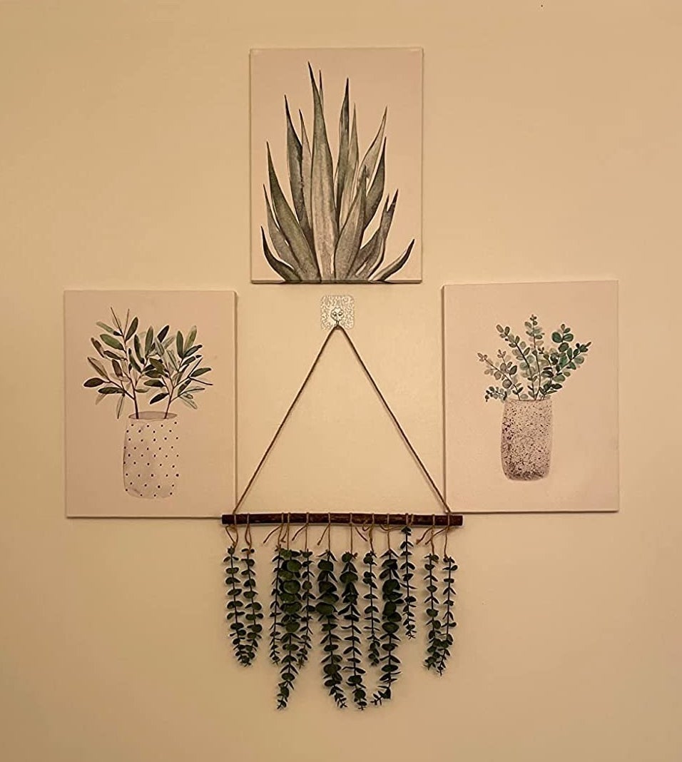 a reviewer photo of three paintings of plants on a white wall with the faux eucalyptus decor hanging below