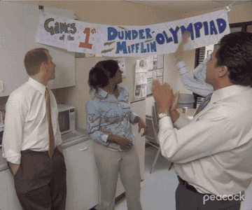 pam gesturing to a sign that says games of the 1st dunder mifflin olympiad on the office