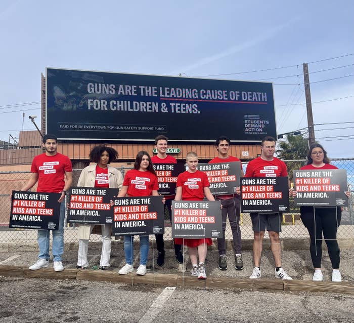 Eight young adults are standing outside beneath a billboard reading "guns are the leading cause of death for children and teens," each of them carrying a sign reading "guns are the number one killer of kids and teens in America"