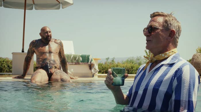 Dave Bautista sits on the edge of a swimming pool and Daniel Craig walks in the pool