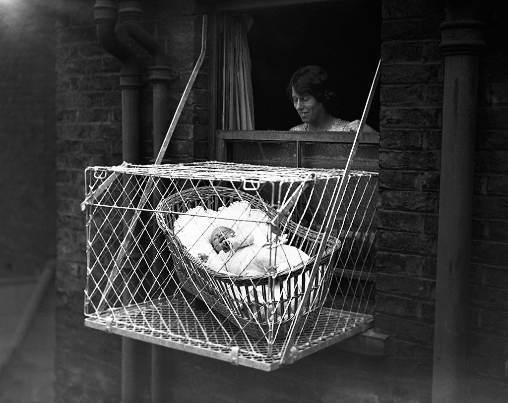 A baby in a crib inside a cage hanging outside a tenement window as a woman looks on
