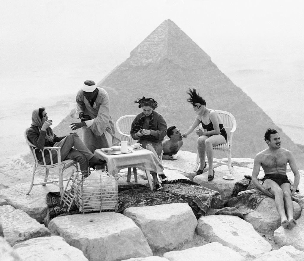 Black-and-white photo of people sitting in chairs around a small table on rocks with a pyramid behind them