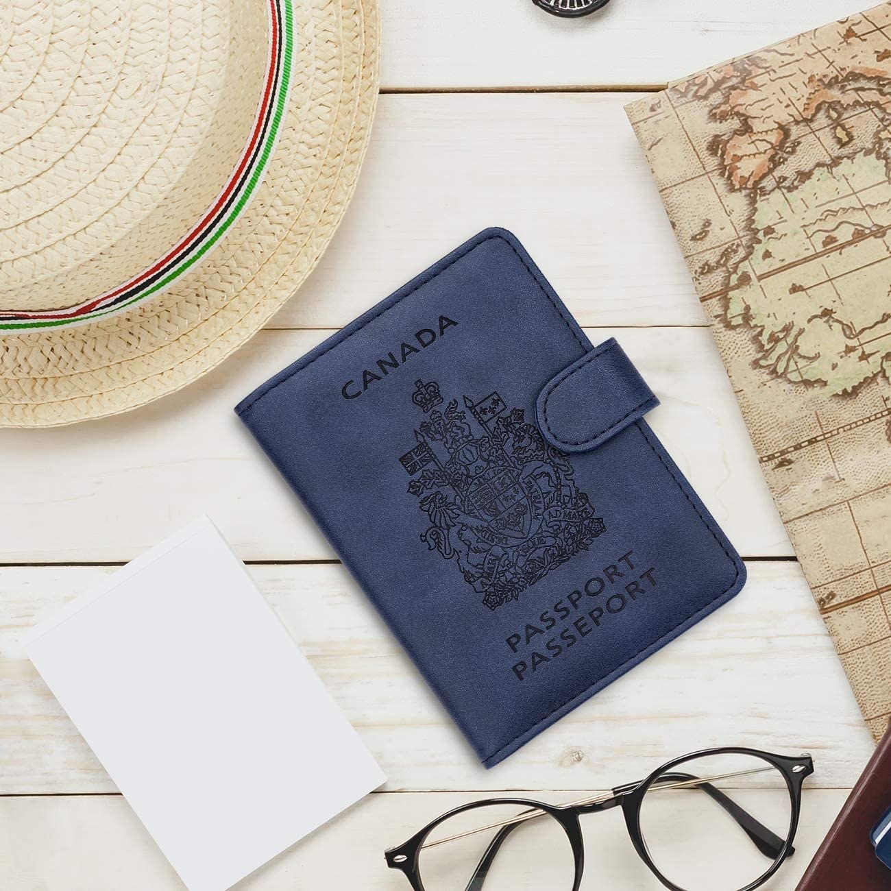 a passport holder that looks like a canadian passport on a wood surface next to a hat, a map, and glasses