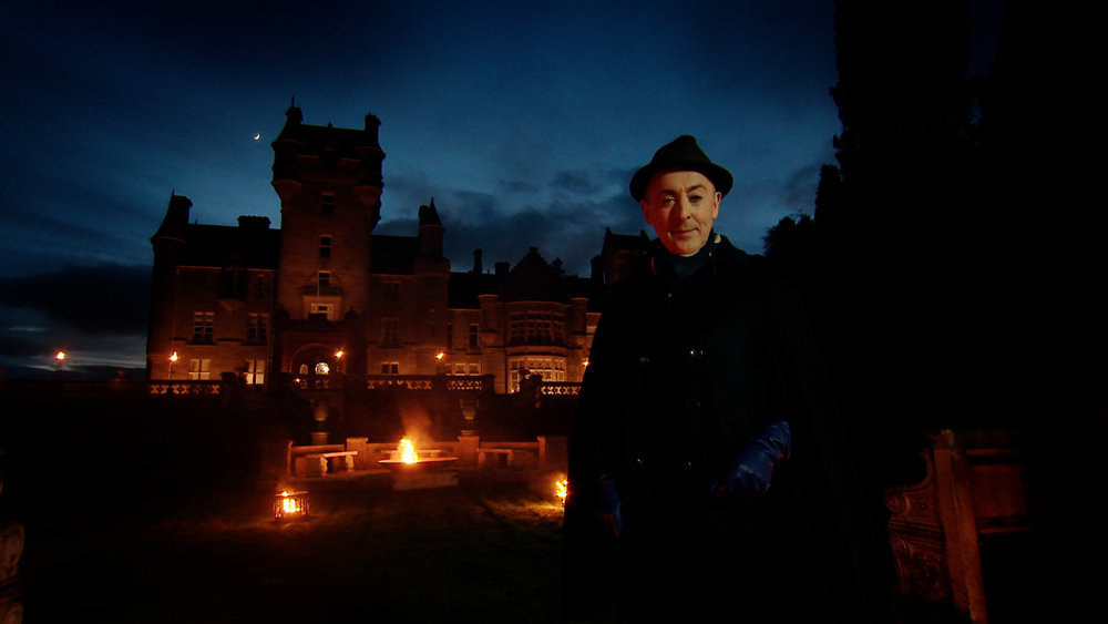 Alan Cumming stands outside of a castle