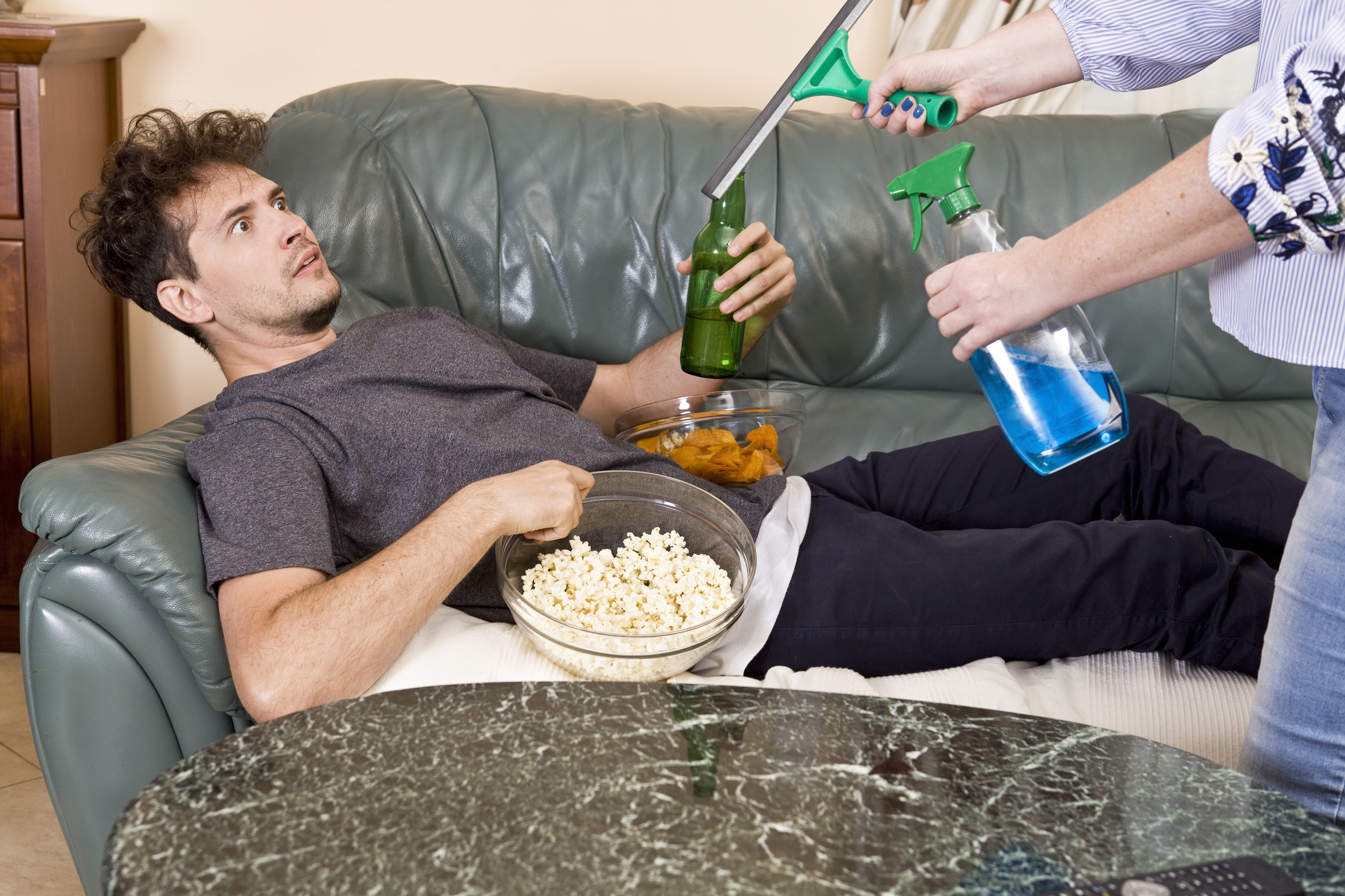 lazy person on couch being bombarded by partner holding cleaning supplies