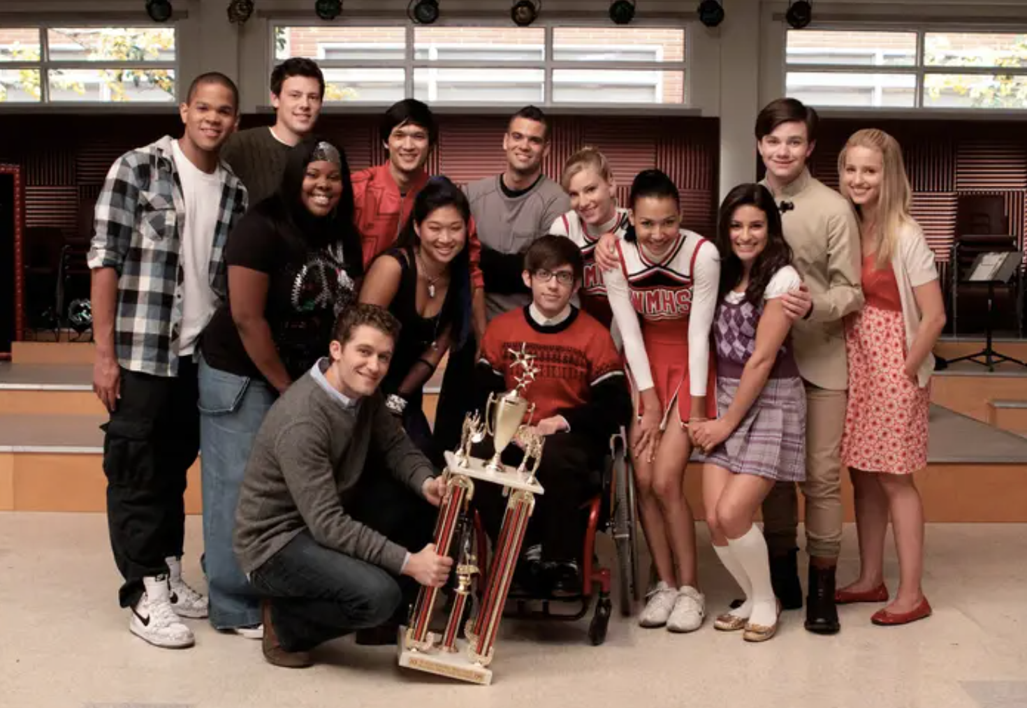 The Glee cast smiles for a group photo with a trophy in a classroom