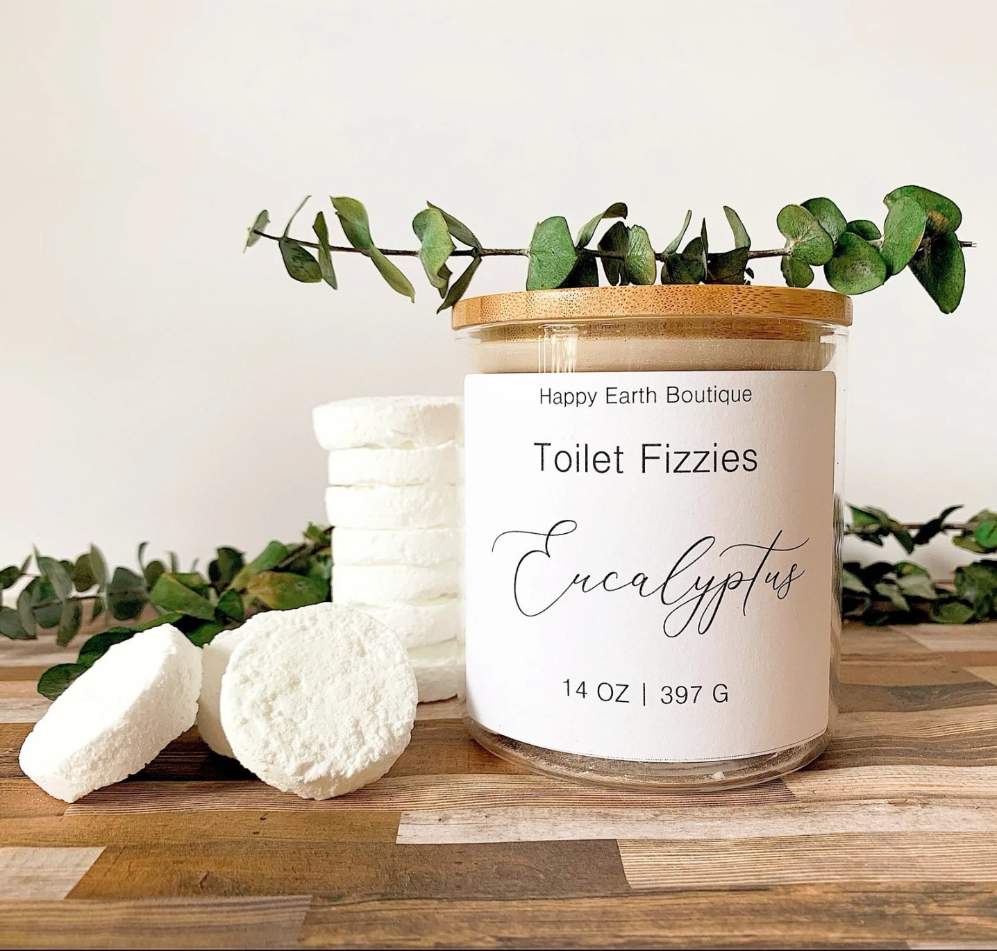 the white jar of toilet fizzies, next to several of the white tablets, on a wood surface with eucalyptus branches