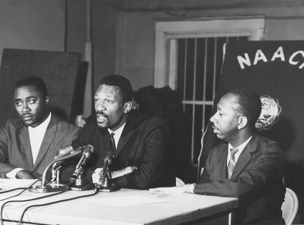 black and white image of three Black men sitting behind microphones