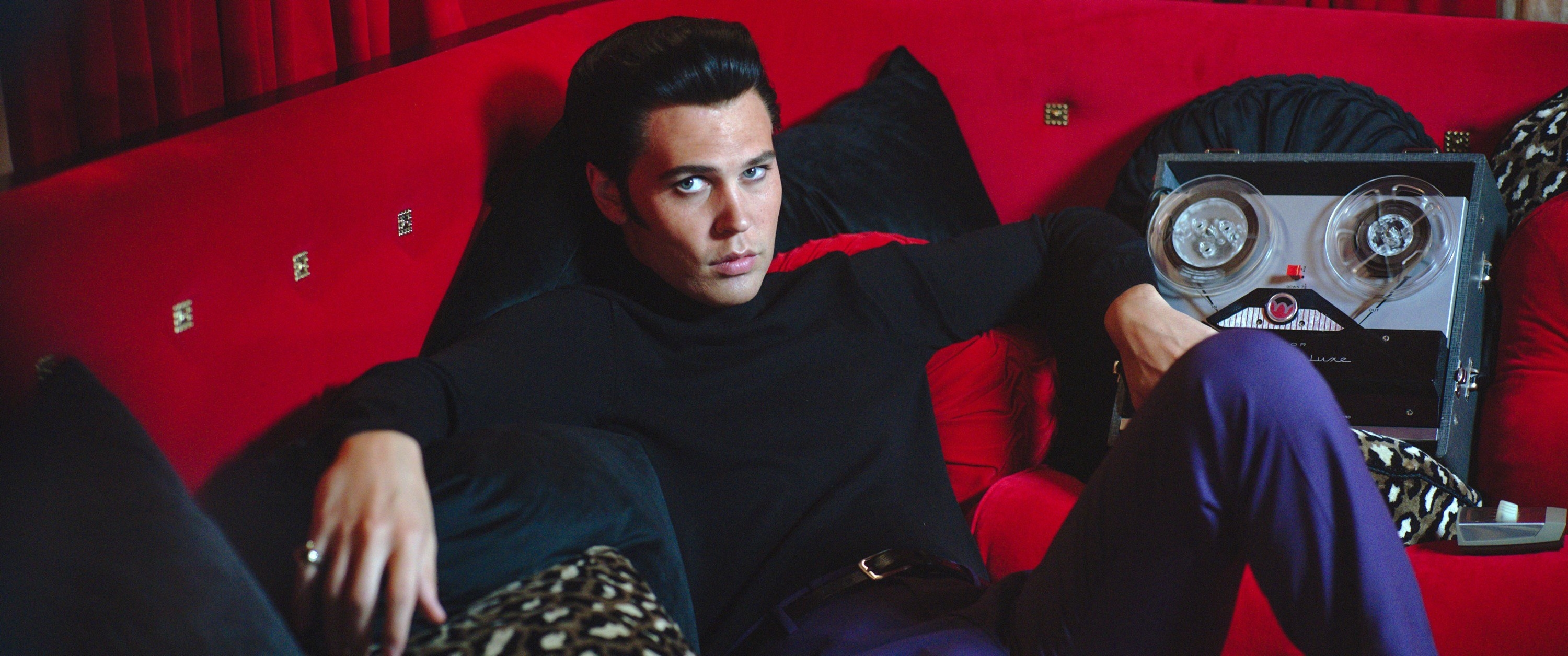 Austin as Elvis lounging on a couch while wearing a turtleneck and slacks