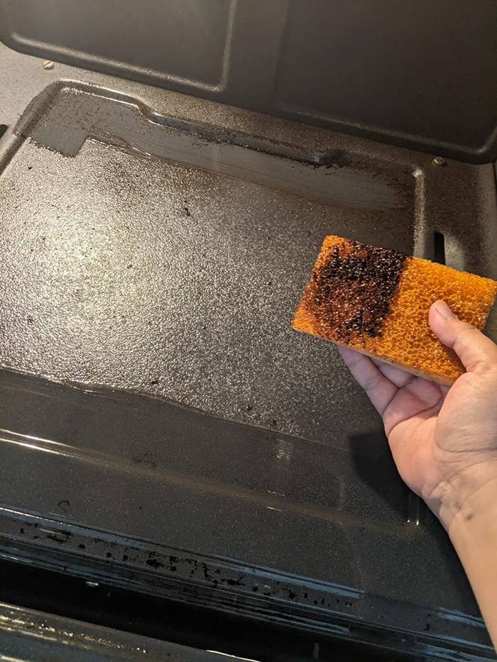 a reviewer photo showing a hand holding an orange sponge covered in black grime. you can see from the oven behind it, that the dirt came off from one swipe of the oven