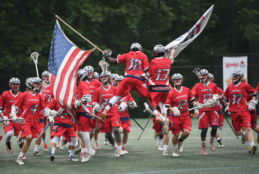 Lacrosse team celebrates with American flag on the field; one player is lifted by teammates