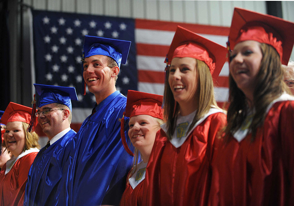 Graduates in caps and gowns smiling during a ceremony, American flag in the background