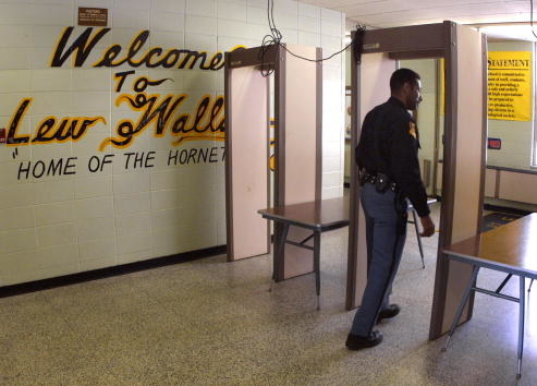 Security personnel walking through metal detectors at a school entrance with welcome sign overhead