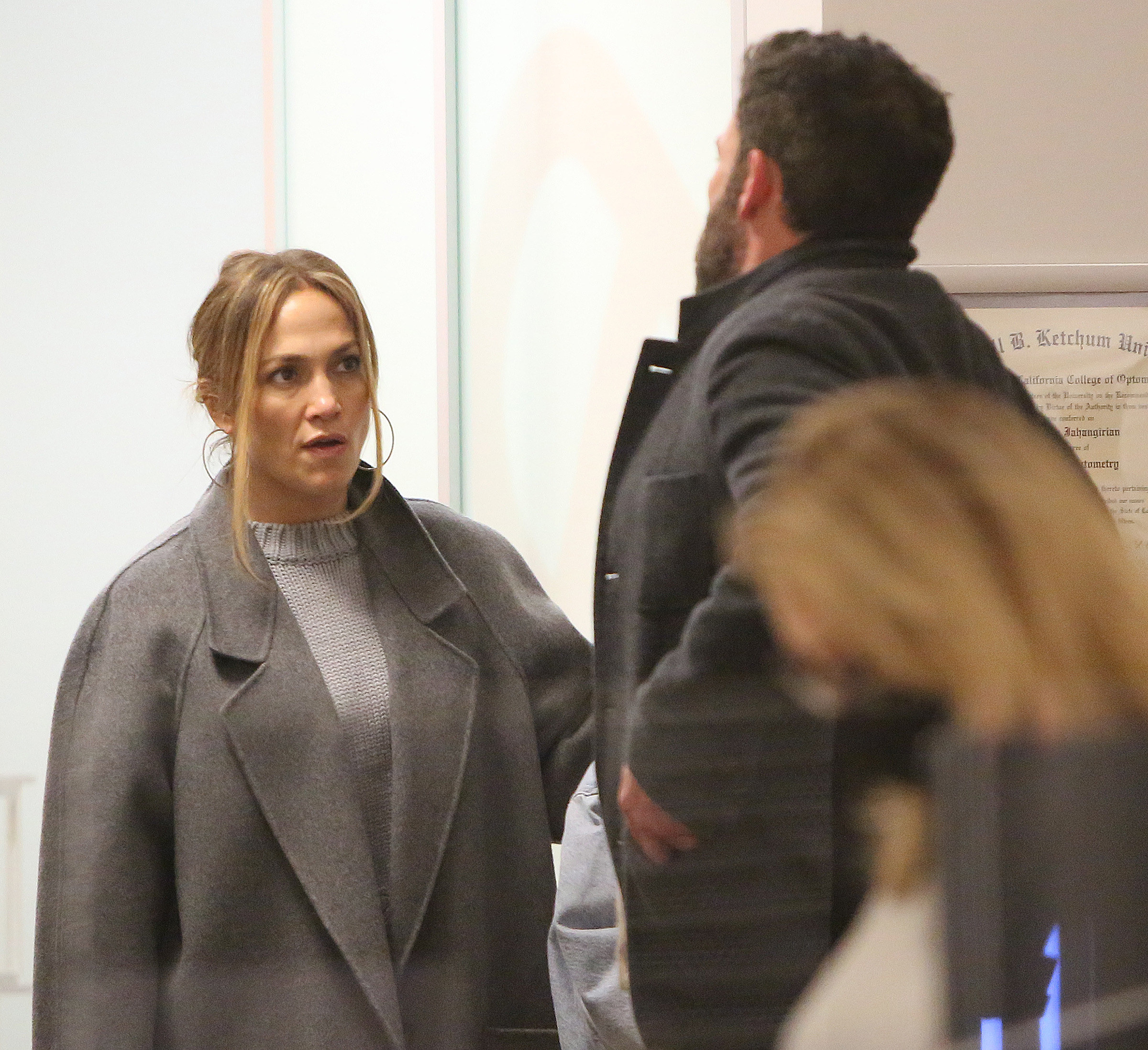 JLo and Ben wearing winter coats and looking at each other while standing inside a building