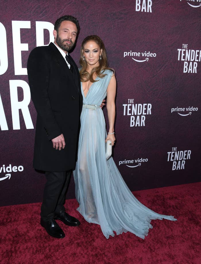 Ben and JLo on the red carpet. nBen is wearing a suit and tie with a long coat. JLo is wearing a flowing Grecian-inspired gown with a plunging neckline