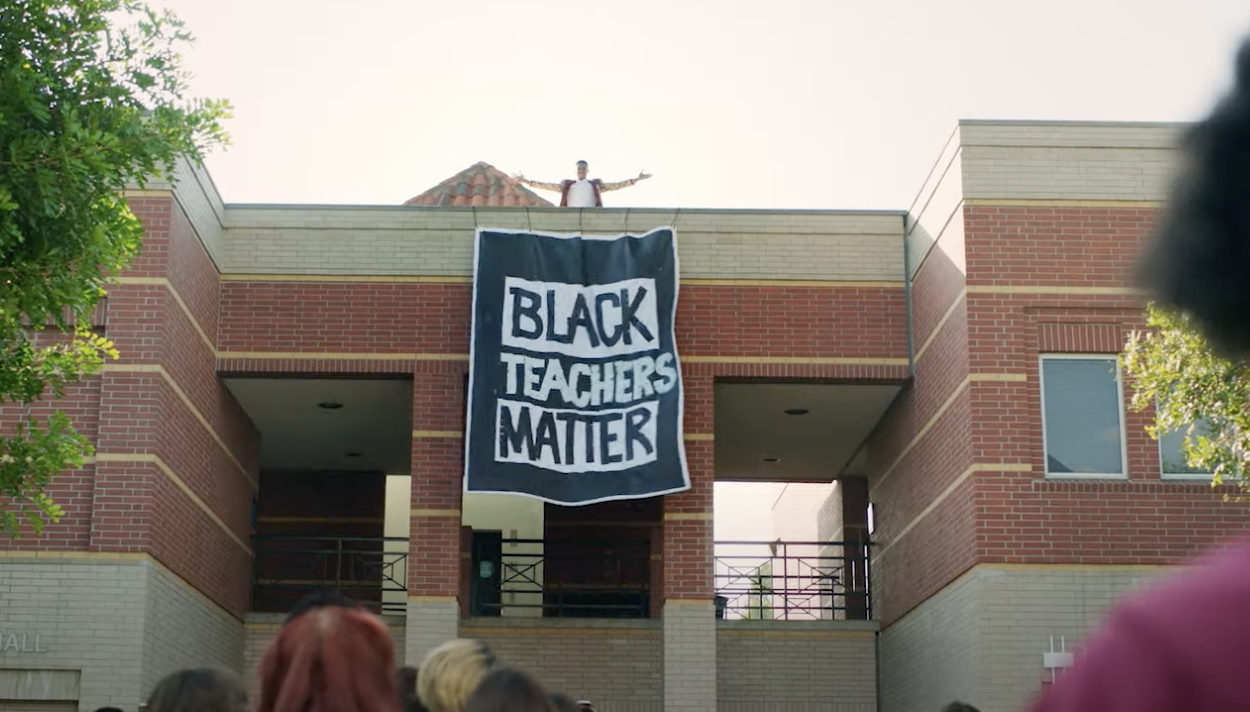 Will unveils a Black Teachers Matter sign from the top of one of the school buildings as people below look on