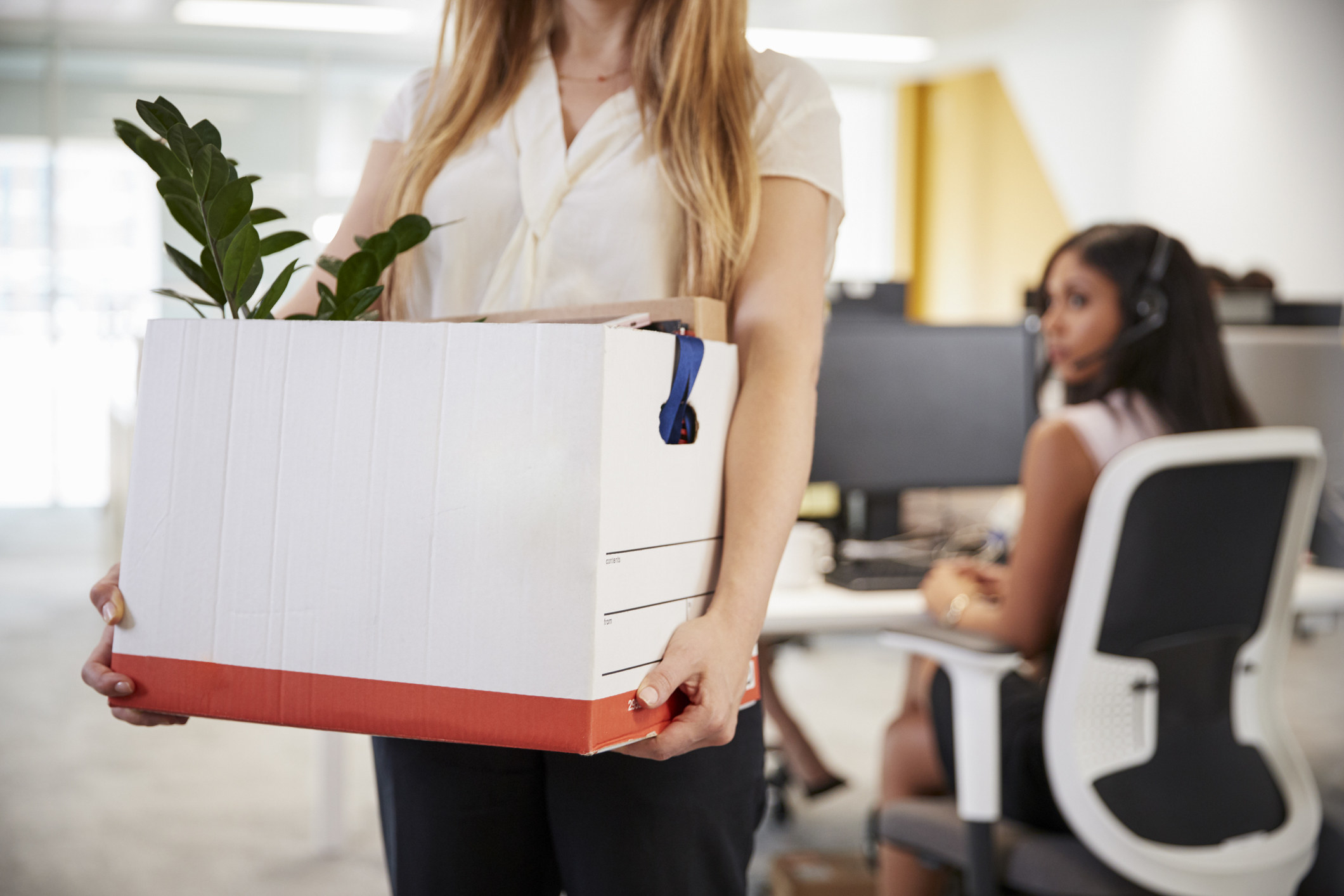 Woman holding a box of office supplies with a plant, indicating she might be leaving her job or relocating desks