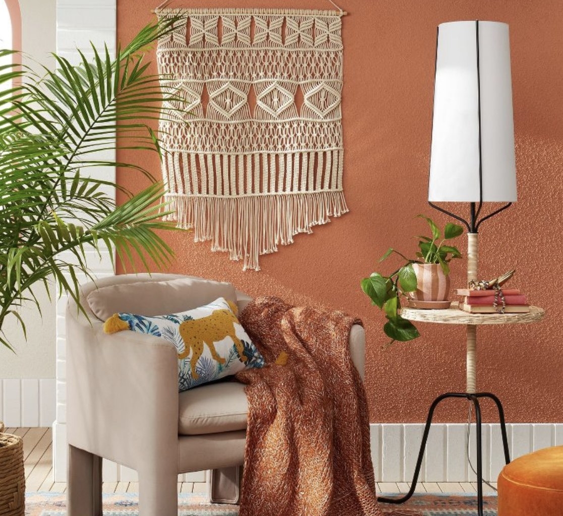 An orange wall with a macrame wall hanging hanging on it with a chair and throw blanket