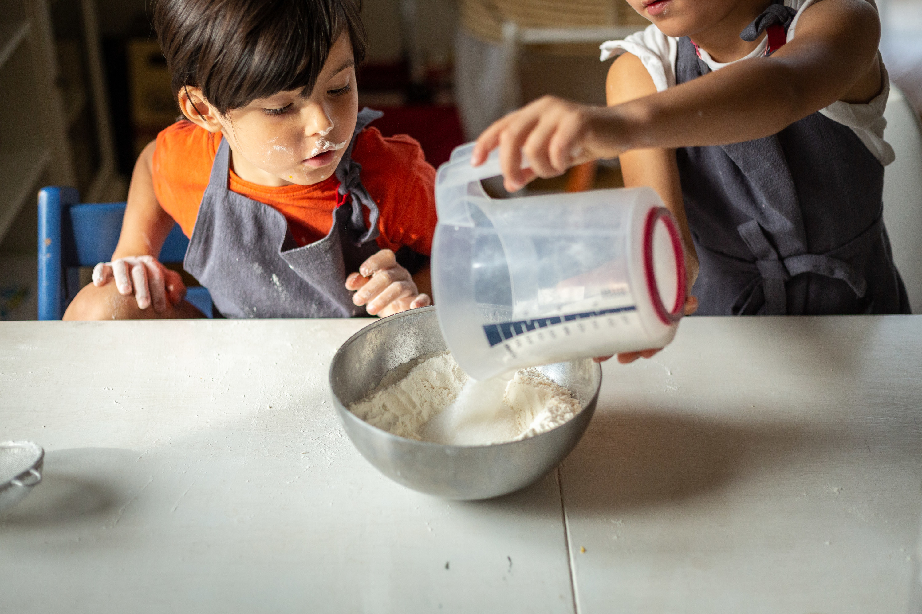two kids wearing grey aprons preparing to bake adding sugar to the flour