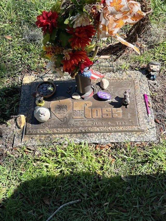 A grave plaque on the ground that says Bob Ross and has an engraving of his face, where fans have left small gifts like stone birds, an american flag, and a Funko pop figurine of Bob