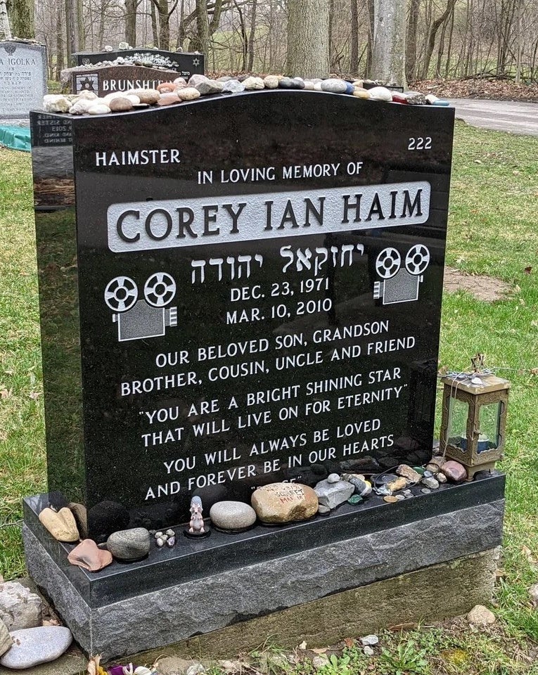 A tombstone with corey&#x27;s name, film cameras, and the text our beloved son, grandson, brother, cousin, uncle, and friend etched on it