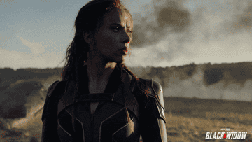 black widow aka scarlett johansson stands in a desert, flames behind her engulfing some structure of some kind