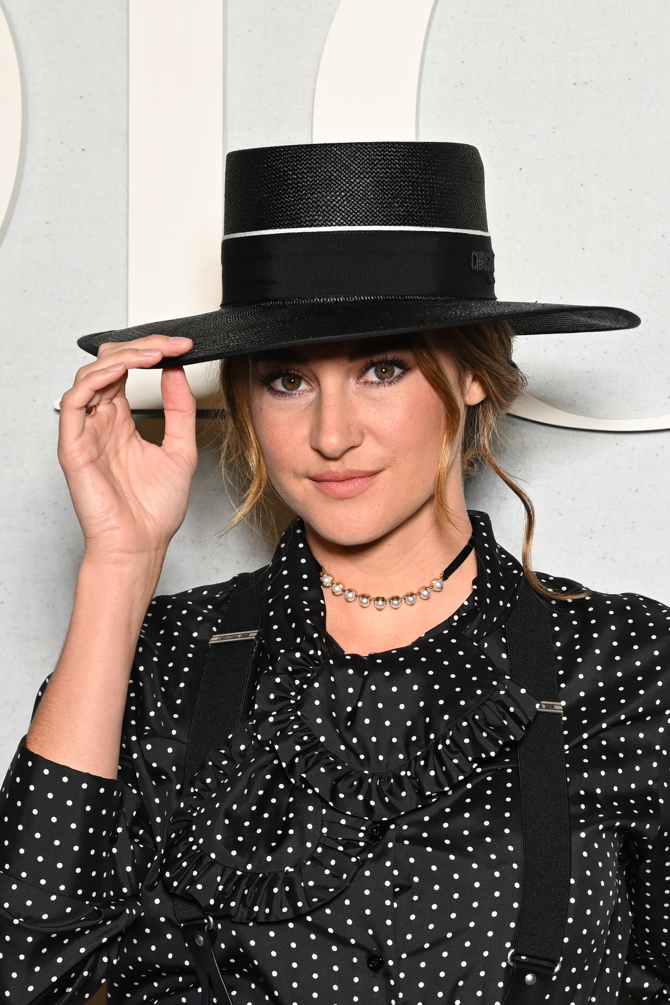 Shailene tipping her wide-brimmed hat as he poses for a photo. Shailene is also wearing a polka dot blouse and suspenders