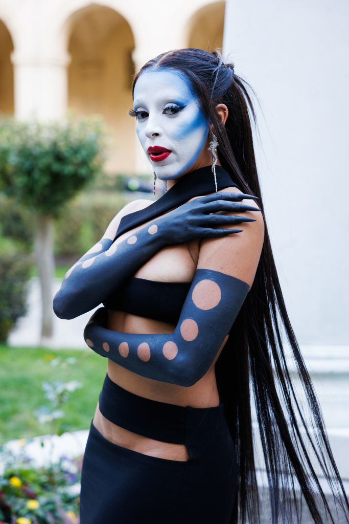 Doja crosses her arms across her body to show off her body paint and long acrylic nails. She also has face paint