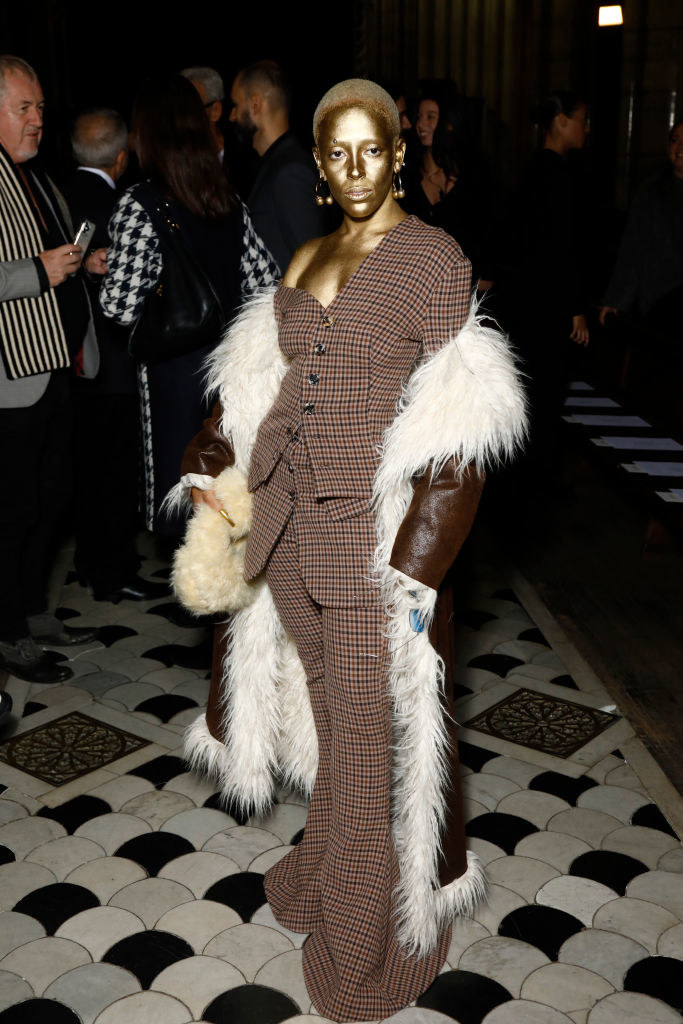 Doja wearing gold face paint, a checkered pantsuit, and a long coat