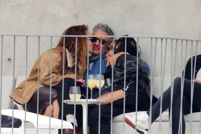 Rita and Emma kissing Taika as they sit on a balcony with drinks on the small table in front of them