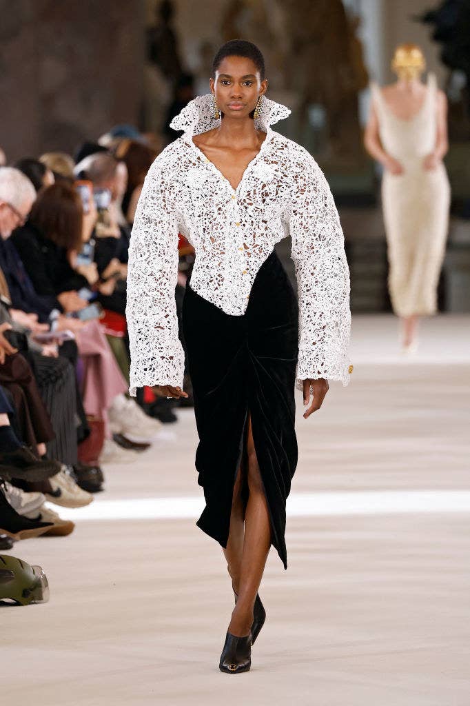 A model wears a dress with a white lace top and popped collar and oversized sleeves and a form-fitting skirt bottom as they walk down the runway