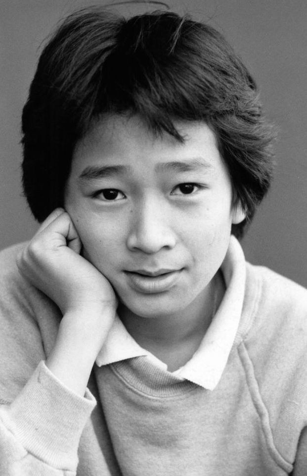A headshot of Quan wearing a sweater and collared shirt as he leans his head in his right hand