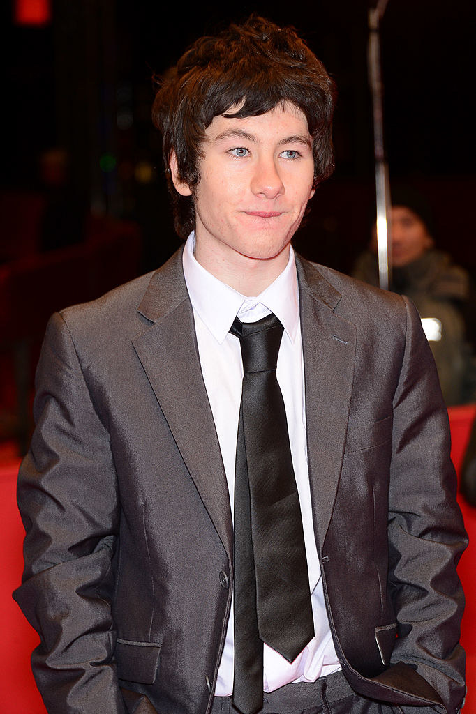 Keoghan in a suit and tie at a red carpet event. He has shaggy hair and is biting his bottom lip