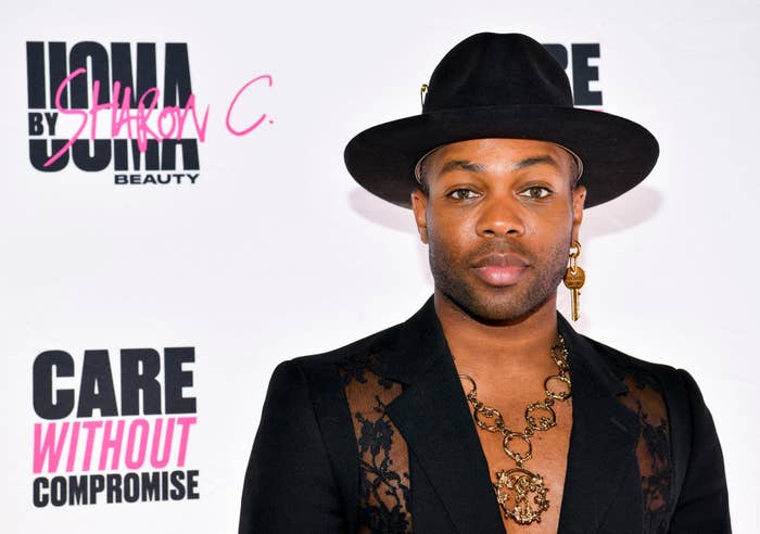 A closeup of Todrick wearing a blazer with lace panels on both sides of the jacket, a fedora, and a single key as an earring on the left ear