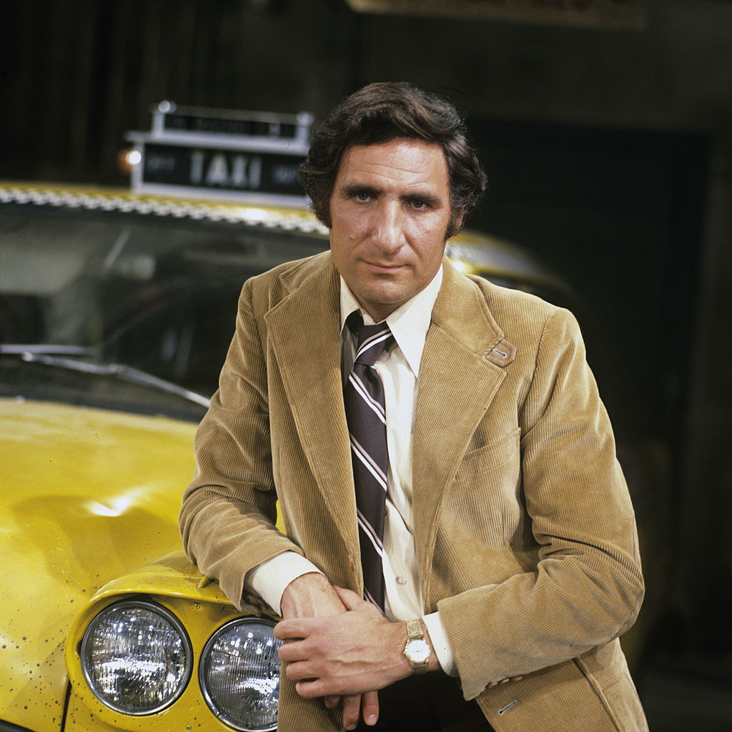 Hirsch leaning against a taxi cab. He is wearing a corduroy jacket, a dress shirt, and tie