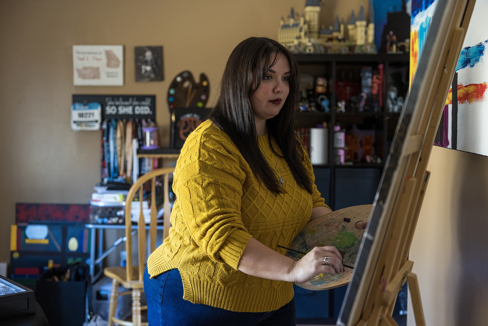 A woman wears a yellow knit sweater and stands with a palette for paint and paintbrush at an easel