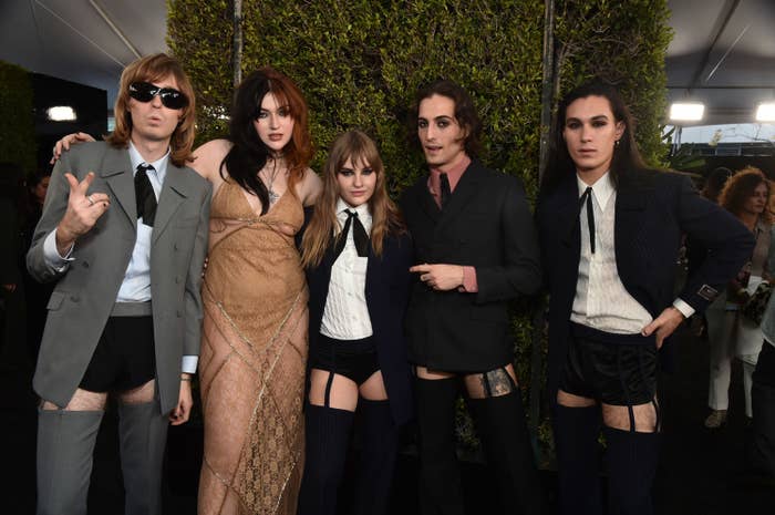 The members of Måneskin posing for a group photo at the American Music Awards. They&#x27;re all wearing suit jackets and ties along with dress shirts and very short shorts and stockings held up by garter belts