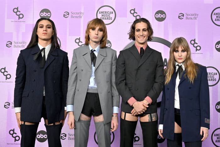 The members of Måneskin posing for a group photo at the American Music Awards. They&#x27;re all wearing suit jackets and ties along with dress shirts and very short shorts and stockings held up by garter belts