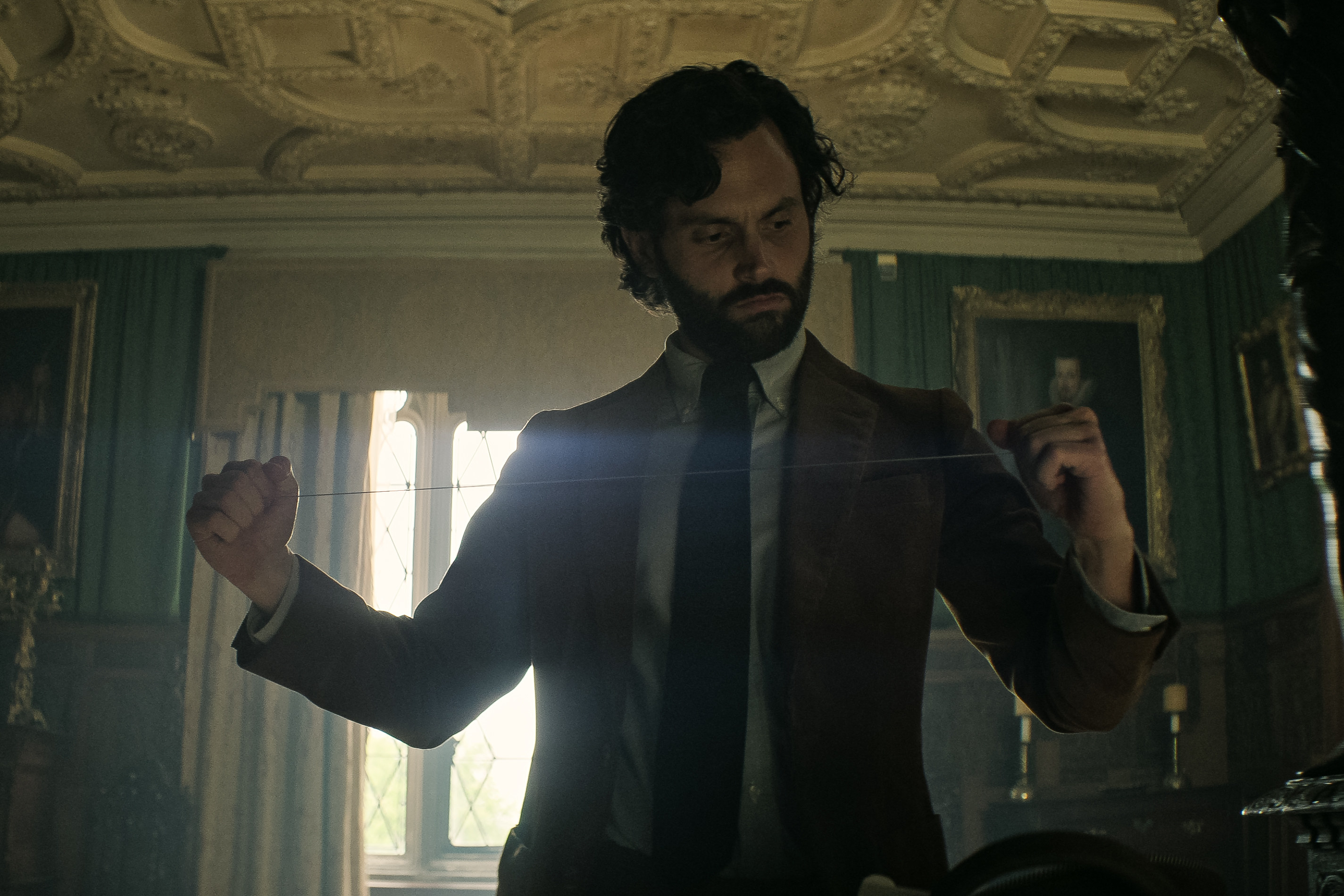 Penn Badgley holding a piece of string while standing in an ornate room in You