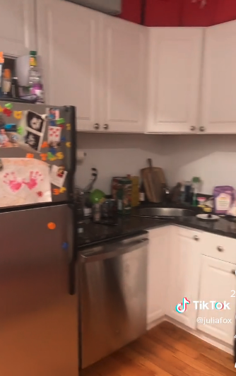 Julia&#x27;s kitchen countertops are filled with everyday culinary essentials like a cutting board and cleaning products. There is a cute painted hand piece of art that her son like made attached to the freezer door