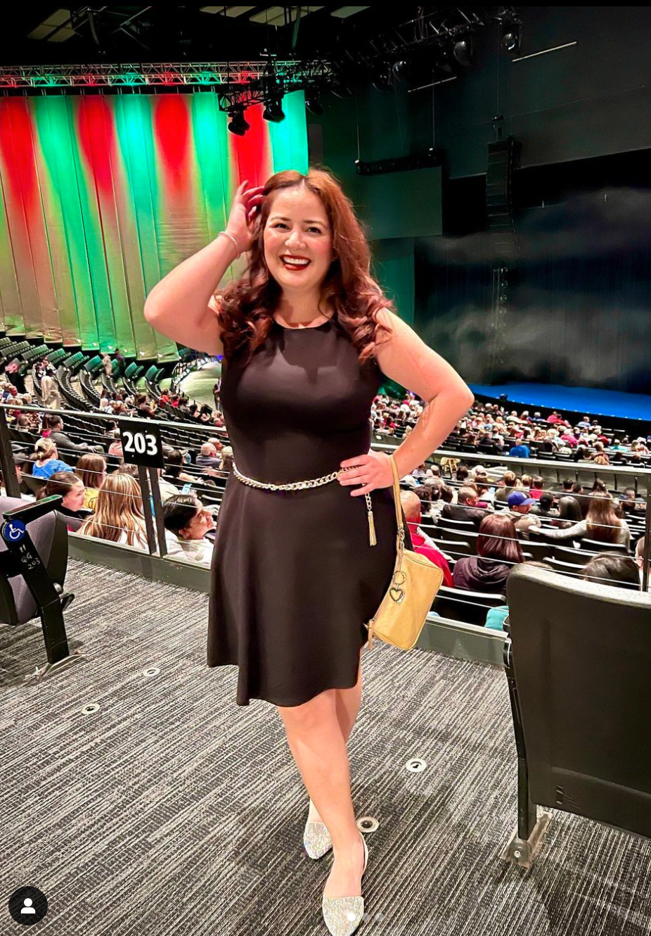 Emily smiles as she poses for a photo a theater while wearing a knee-length sheath dress with a chain belt and sequined flats