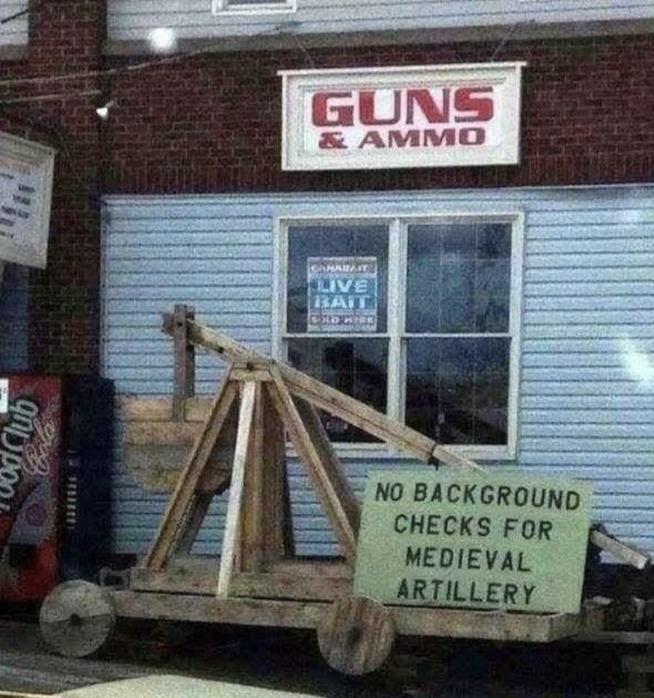&quot;No background checks for medieval artillery&quot; sign on an ancient military device for hurling missiles outside a &quot;Guns &amp;amp; Ammo&quot; store