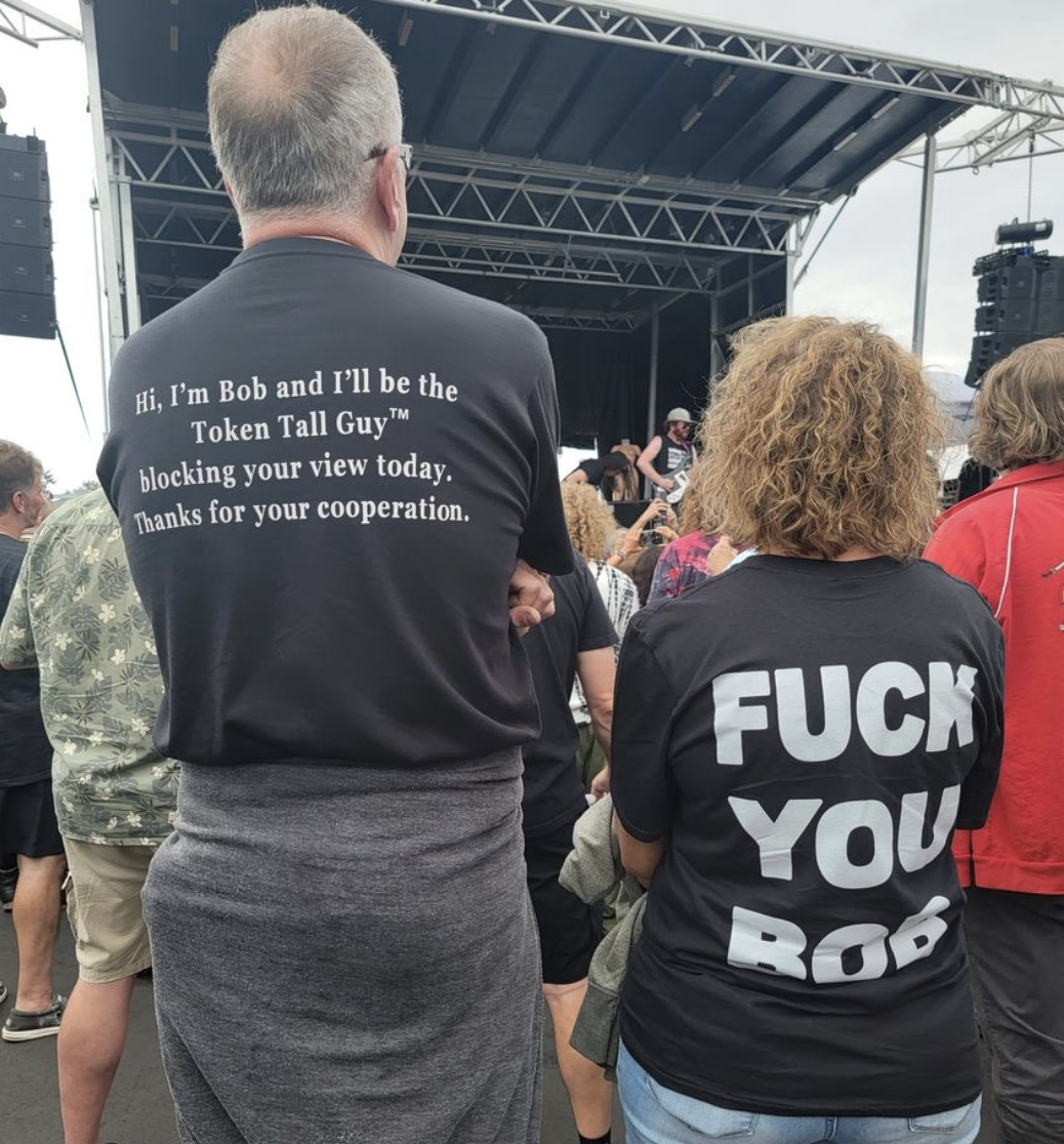 Man with &quot;Hi, I&#x27;m Bob and I&#x27;ll be Token Tall Guy blocking your view today, thanks for your cooperation&quot; and a woman with &quot;Fuck you Bob&quot; on the backs of their T-shirts