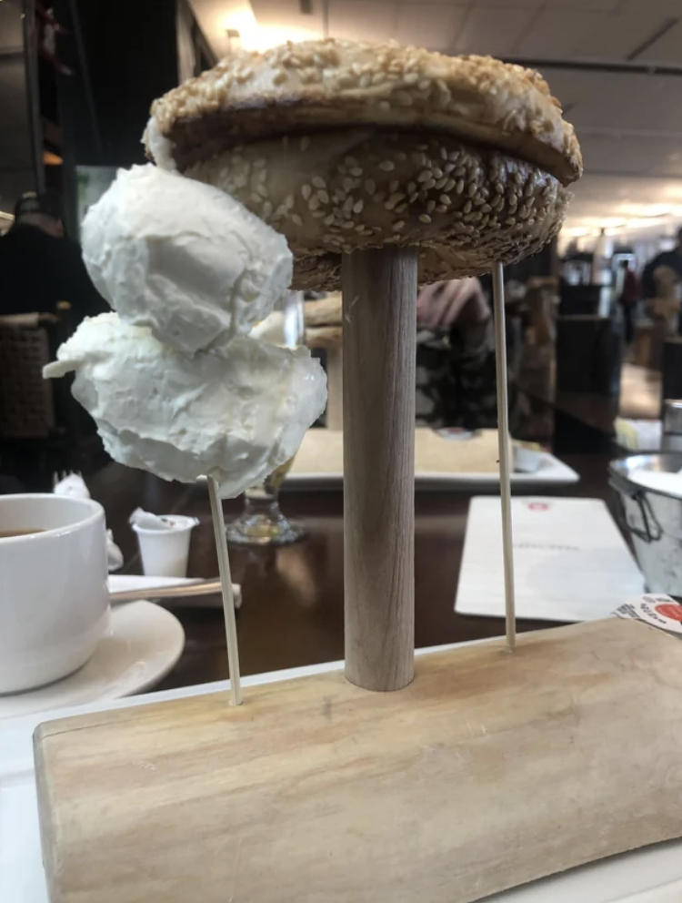 A sesame bagel stuck on some kind of rod attached to a cutting board, and cream cheese on a stick