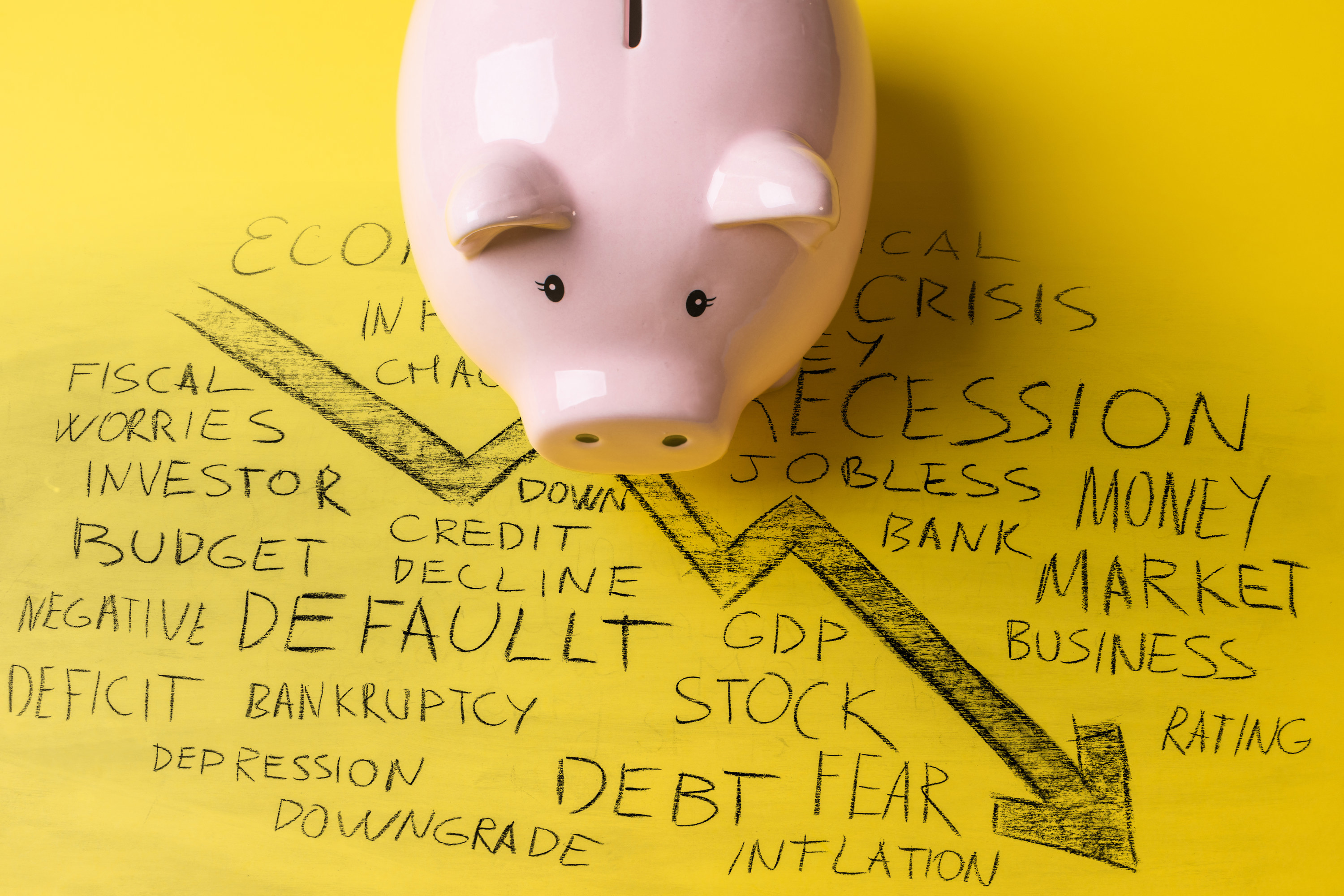 A piggy bank on a surface that has a stock market arrow going down surrounded by handwritten words like debt fear inflation worries negative jobless and downgrade
